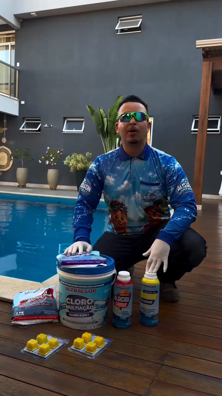 Alisson agachado na beira de uma piscina, de uniforme azul e luvas, com baldes de cloro e frascos de algicida Ultraclor